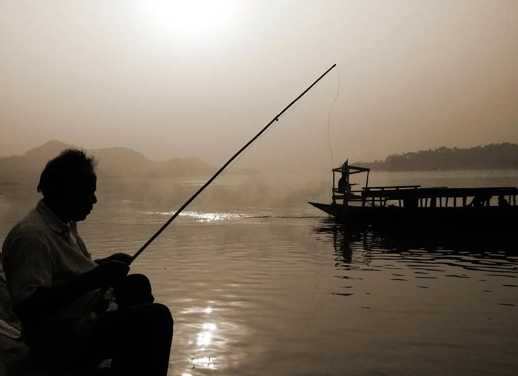A silhouetted fisherman with a long rod at the water’s edge in soft evening light